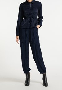 Navy blauwe jumpsuit met een ritssluiting aan de voorkant, kraaghals, elastische manchetten en twee voorzakken. Draag met zwarte enkellaarsjes.