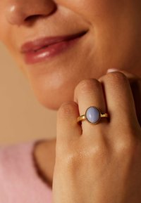 Gouden ring met een in zetting geplaatste ovale lichtblauwe steen, met een gestructureerde band, gedragen op een hand tegen een zachte achtergrond.