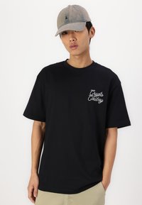 Sort T-shirt med korte ærmer og en afslappet pasform, med hvid broderet tekst på venstre bryst. Bæres med en lys farvet kasket.