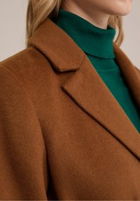 Bruine wollen jas met een kraag met inkeping en zachte textuur, gedragen over een groene coltrui. Close-up van de stofdetails.