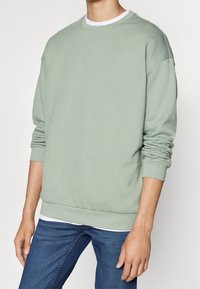 Grön bomullströja med rund halsringning, ribbade ärmslut och kant, avslappnad passform, lager över en vit skjorta, tillsammans med blå jeans.