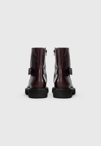 Bottines en cuir bordeaux avec un bout rond, semelle noire épaisse, fermetures éclair latérales et une sangle décorative à boucle autour de la cheville.