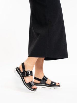 Füße in schwarzen Lackledersandalen mit Schnallen und weißen dicken Sohlen, getragen zu einem langen schwarzen Rock.