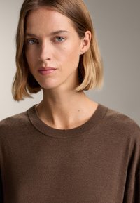 Barna kötött pulóver, kerek nyakkivágással és bordázott aljjal. Az anyag puha, sima tapintású, és kissé laza szabású.