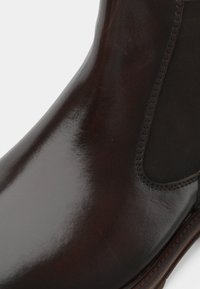 Bota de tobillo de cuero marrón con un acabado brillante, paneles laterales elásticos y una suela resistente. Presenta detalles de costura a lo largo de los bordes.