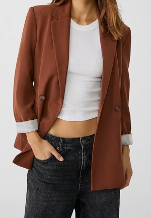 Frau trägt rostfarbenen Blazer über weißem, kurz geschnittenem, geripptem Top mit hochgekrempelten Ärmeln, kombiniert mit hoch taillierter schwarzer Jeans, Hand in der Tasche.