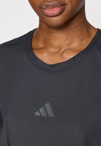 Μαύρο αθλητικό πουκάμισο με υφή υφάσματος, στρογγυλεμένο λαιμόκοψη και διακριτικό λογότυπο Adidas σε πιο σκούρο τόνο στο μπροστινό μέρος.