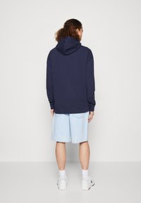 Dunkelblaue Kapuzenjacke mit großer Kapuze, gerippten Bündchen und Saum, kombiniert mit hellblauen Jeansshorts, die bis knapp über das Knie reichen. Weiße Sneakers mit Socken.