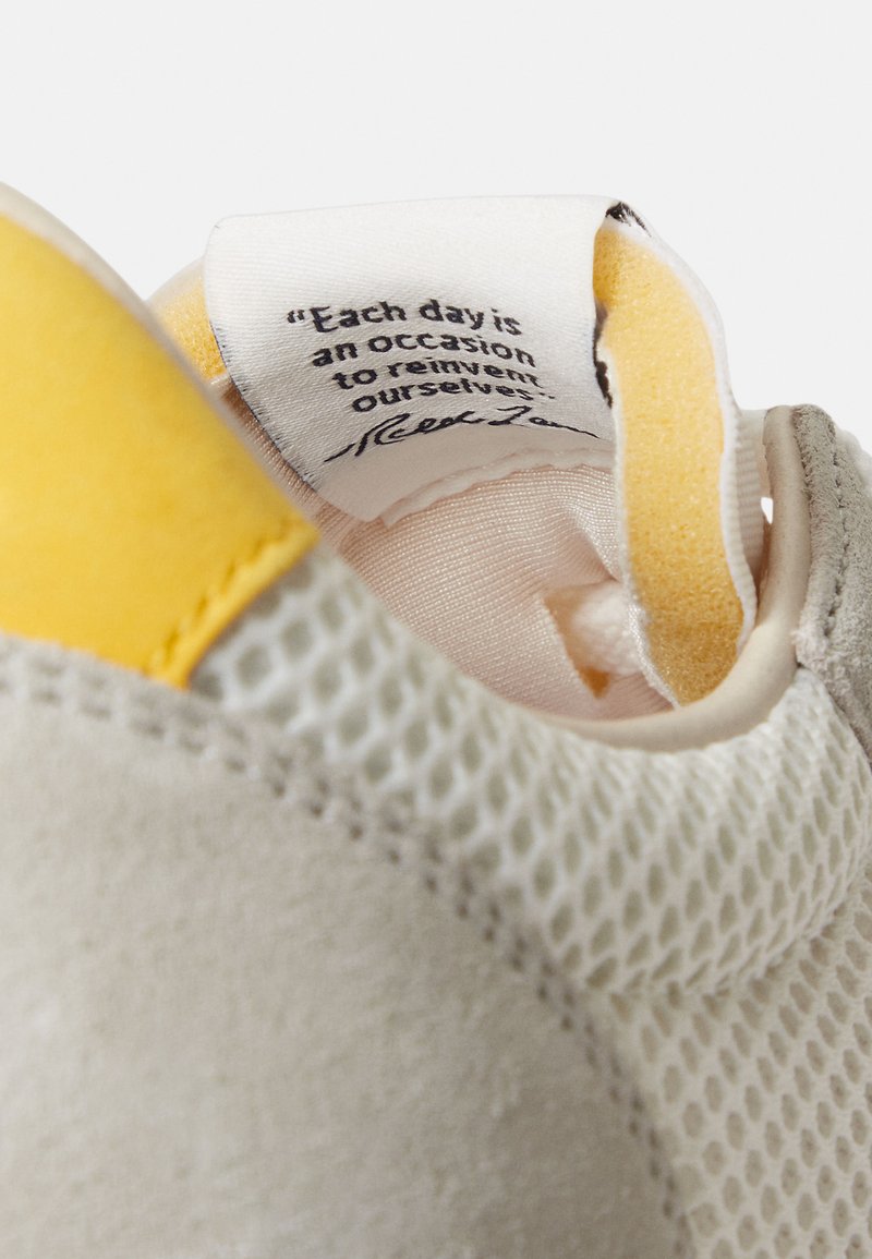 Baskets gris clair avec un accent jaune, dotées d'une tige en mesh et en daim. Comprend une étiquette à l'intérieur avec une citation motivante.