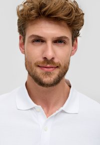 Witte polo met een gestructureerde stof, voorzien van een knoopsluiting met twee knopen en een klassieke kraag. Model met kort, tousled haar en een baard.