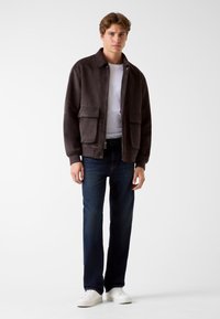 Giacca in suede marrone con due tasche frontali, chiusura con zip e colletto, abbinata a jeans scuri e sneakers bianche.