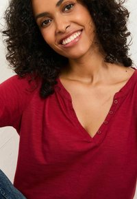 Lächelnde Frau mit lockigem, dunklem Haar, die ein rotes, knöpfbares V-Ausschnitt-Shirt und blaue Jeans trägt, vor einem hellen Hintergrund.