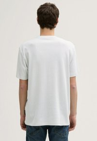 Tricou alb cu mânecă scurtă, dintr-un material texturat, cu guler rotund și un eticheta în partea din stânga jos. Vedere din spate, purtat cu blugi albaștri.