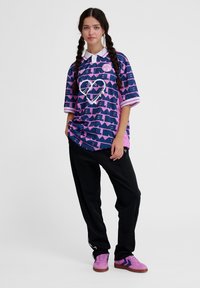 Camisa polo oversized em azul marinho e rosa com padrões geométricos e um design de coração, combinada com calças pretas e ténis rosas com detalhes em preto.