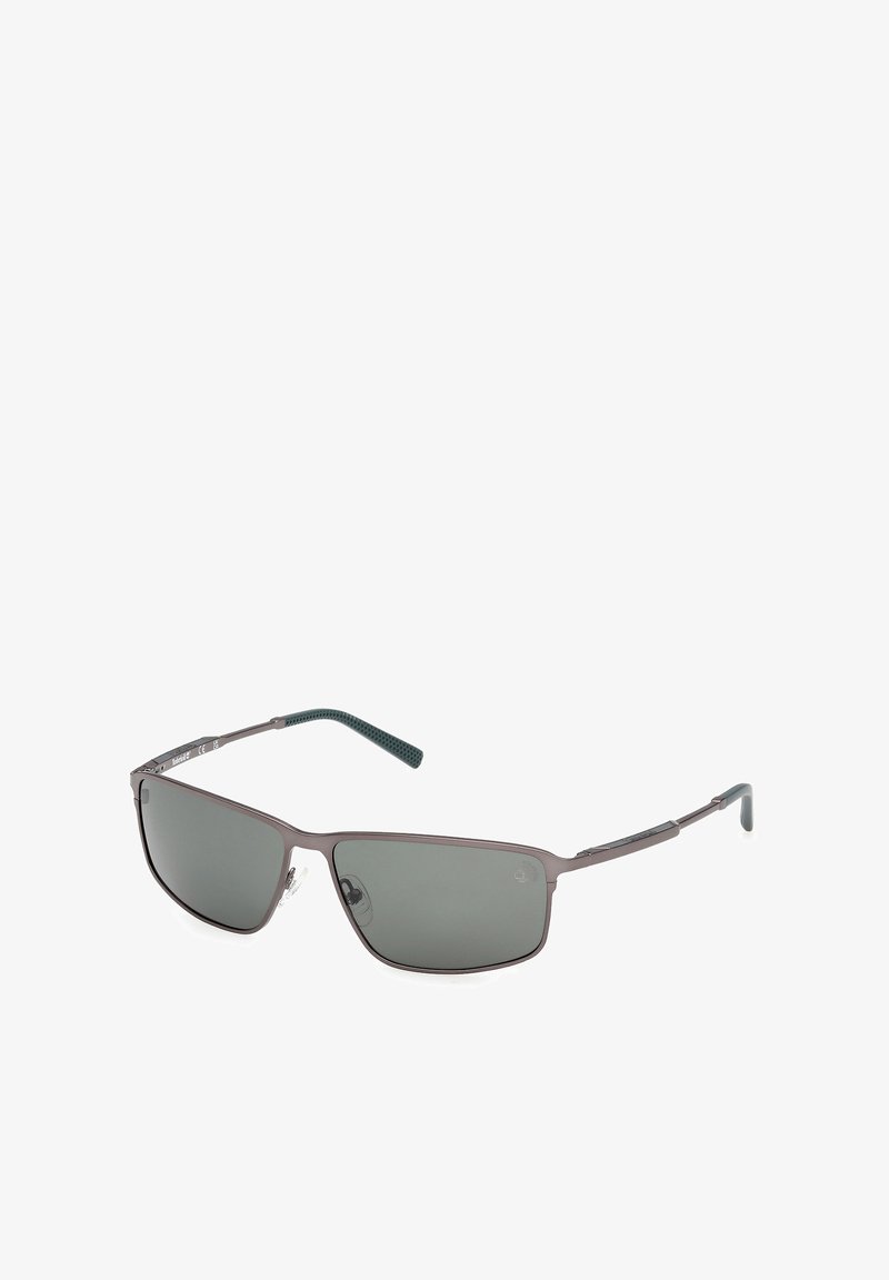 Lunettes de soleil à monture rectangulaire en métal gris avec verres teintés foncés et embouts de branche noirs texturés, inclinées sur fond blanc.