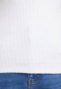 Bílý ribovaný top s vertikálními pruhy, který má těsný střih. Kombinovaný s modrými denimovými džíny s detaily šití.