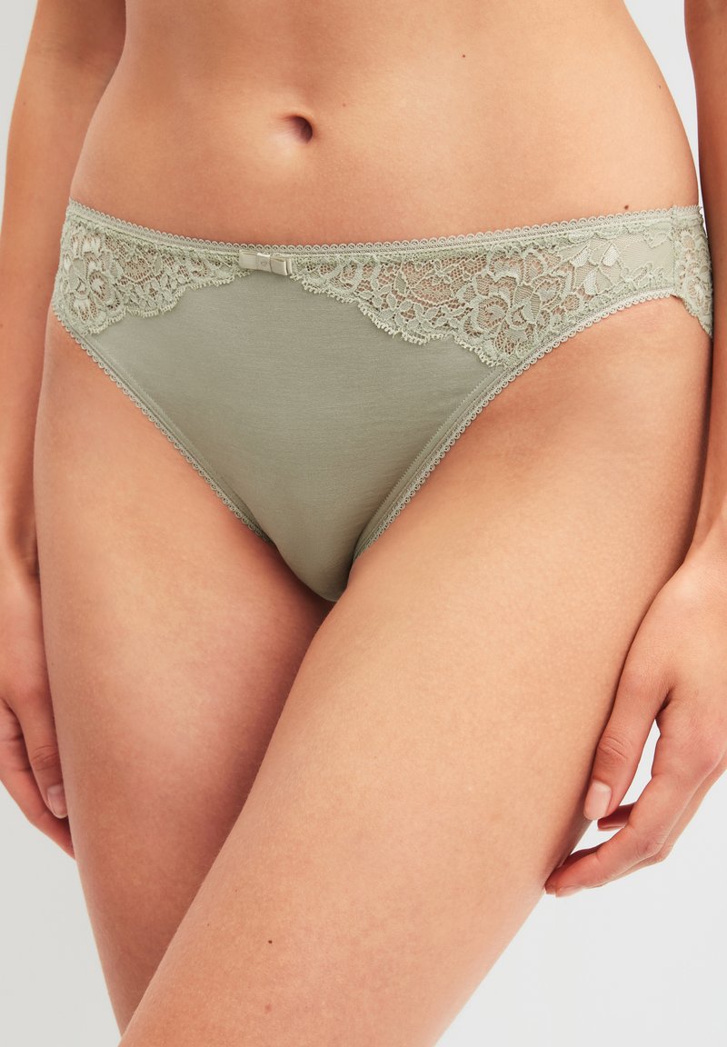 Femme portant un bikini en coton vert clair avec bordure en dentelle, visible de la taille jusqu'aux cuisses, la main posée sur la cuisse.
