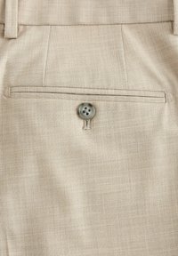 Gros plan sur un tissu beige montrant une poche arrière de pantalon avec une fente horizontale et un seul bouton gris cousu sous la poche.