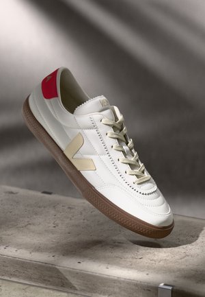 White leather sneaker with beige accents, red heel, textured brown rubber sole, and contrasting stitching. Laces positioned centrally.