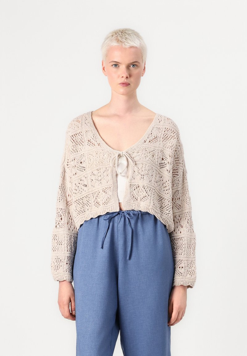 Strikket cardigan i lys beige med åpen heklemønster og knyting foran. Kombinert med løse, lyseblå bukser, som har strikk i livet.