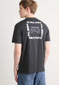 Γκρίζο βαμβακερό t-shirt με λευκή εκτύπωση του λογότυπου "Calvin Klein" και λεπτομέρειες διεύθυνσης στην πλάτη, με κλασικό λαιμόκοψη και κοντά μανίκια.