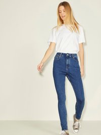 Jeans de mezclilla azul de cintura alta con un corte ajustado, cinco bolsillos, cremallera y combinados con una camiseta blanca sencilla y zapatillas grises.