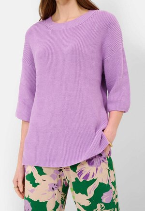 Femme portant un pull en maille lavande avec des manches jusqu'au coude et un pantalon beige avec des motifs floraux verts et violets.