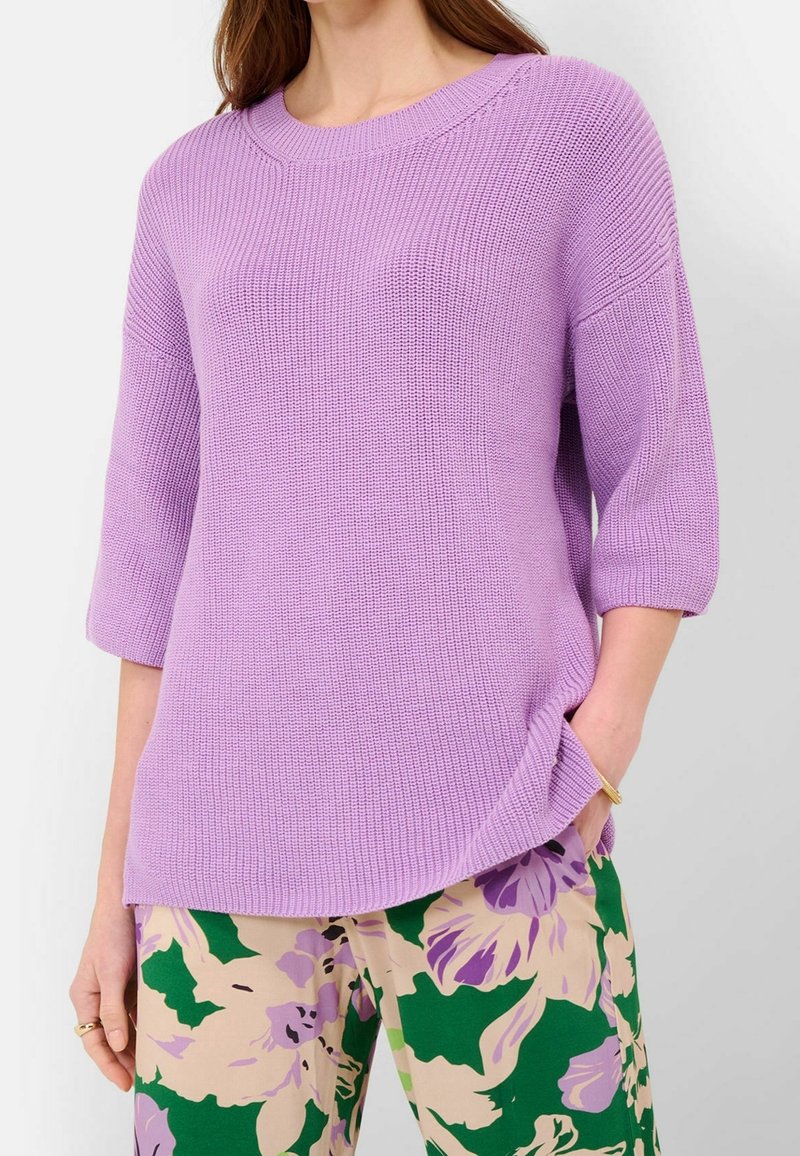 Femme portant un pull en maille lavande avec des manches jusqu'au coude et un pantalon beige avec des motifs floraux verts et violets.