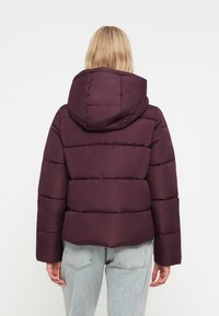 Chaqueta de puffer morada con capucha, que presenta un diseño acolchado y puños elásticos. La chaqueta tiene un corte corto y está hecha de material suave.
