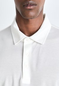 Bílá polo košile s límečkem, vyrobená z texturované látky, s třemi knoflíky a jemnými detaily šití.
