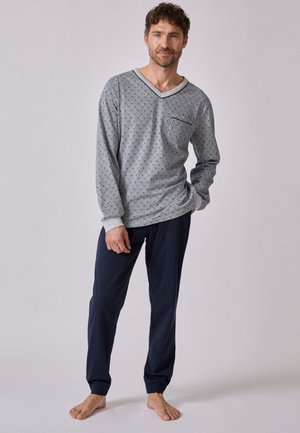 Man staat op blote voeten, draagt een grijs longsleeve met klein patroon en donkerblauwe pyjamabroek tegen een neutrale achtergrond.
