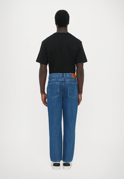 Μπλε τζιν παντελόνι με χαλαρή εφαρμογή, πίσω τσέπες και ένα καφέ δερμάτινο λογότυπο στη μέση. Συνδυασμένο με ένα απλό μαύρο T-shirt.