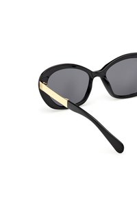 Gafas de sol negras con forma ovalada y lentes oscuros. Presentan un acento dorado en las patillas. Hechas de plástico con una textura suave.