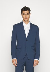 Navy blue suit jacket with notch lapel, single button closure, and two front pockets. Worn over a white button-up shirt. Smooth fabric texture.