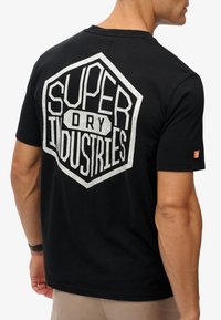 Camiseta de algodón negra con un gran logo gráfico blanco en la parte posterior que dice "SUPER DRY INDUSTRIES", con mangas cortas y cuello redondo.