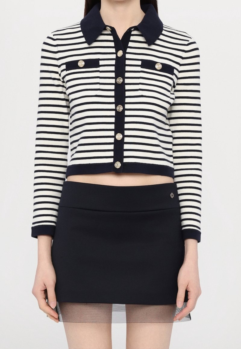 Striped knit cropped sweater in navy and white with a collared neckline, gold buttons, and two front pockets; paired with a navy mini skirt.