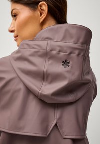Chaqueta impermeable de color malva claro con cuello alto, costuras visibles y un logo destacado en la parte posterior. Textura suave, diseño moderno.