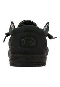 Zapato negro de tela tipo slip-on con un upper texturizado, suela flexible negra con detalles moteados y una etiqueta con el logo "HEY DUDE" en la parte trasera.
