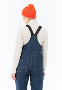 Vrouw met een oranje gebreide beanie, beige ribbeltrui met col, en blauwe denim overall met zwarte banden, gezien van achteren op een witte achtergrond.