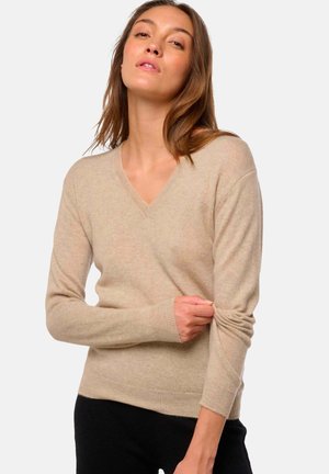 Femme ajustant la manche d'un pull beige à col en V, portant un pantalon noir, devant un fond clair.
