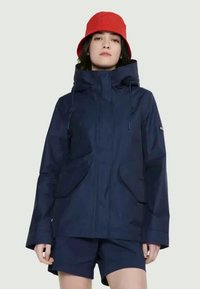 Veste imperméable bleu marine avec capuche, dotée de poches avant, de cordons de serrage ajustables et d'une coupe décontractée, portée avec un short bleu marine et un chapeau seau rouge.