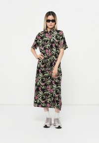 Robe mi-longue florale noire avec manches courtes, feuilles vertes et fleurs roses ; nouée à la taille. Portée avec des baskets grises et des chaussettes blanches.