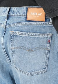 Jeans din denim de culoare albastru deschis, dotate cu o etichetă din piele maro cu brand, un singur buzunar la spate cu cusături curbate și un mic detaliu cu logo roșu.