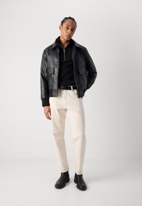 Chaqueta de cuero negra con bolsillos abotonados, suéter negro ajustado y pantalones beige claros, combinados con botas de tobillo negras. Diseño minimalista.