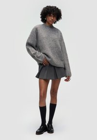 Grauer oversized Pullover mit Rundhalsausschnitt und weicher Textur. Kombiniert mit einem grauen Plisseerock, schwarzen knielangen Socken und schwarzen Loafers.