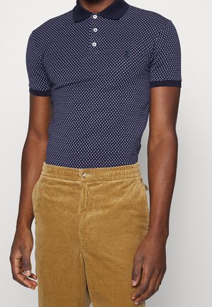 Man die een marineblauw poloshirt met korte mouwen en patroon en mosterdgele corduroy broek draagt, staat tegen een effen achtergrond.