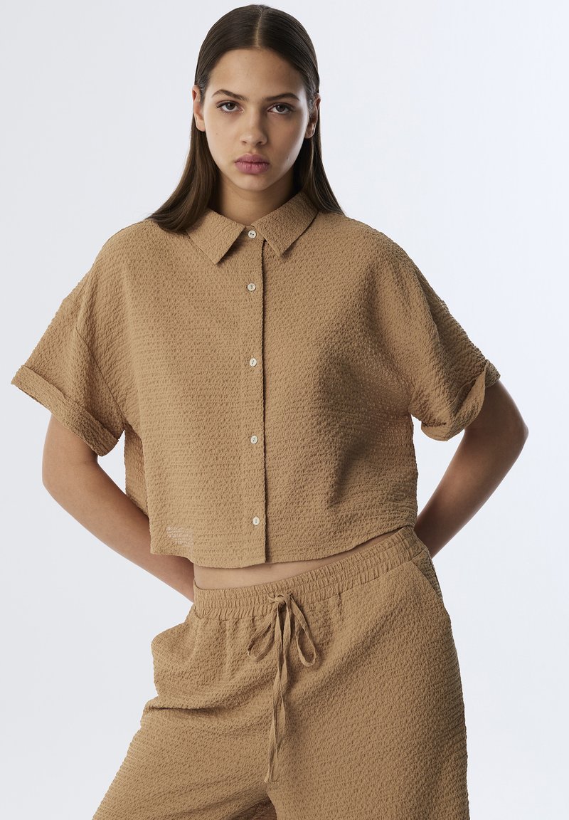 Camiseta de manga corta y recortada en tejido beige texturizado con un cierre de botones en la parte delantera y un cuello puntiagudo, acompañada de pantalones cortos con cintura elástica a juego.