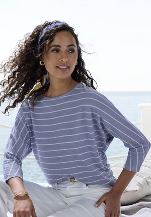 Frau mit lockigem Haar, die ein blau-weiß gestreiftes Oberteil, weiße Hose und ein blaues Kopftuch trägt und an einem sonnigen Tag am Meer sitzt.