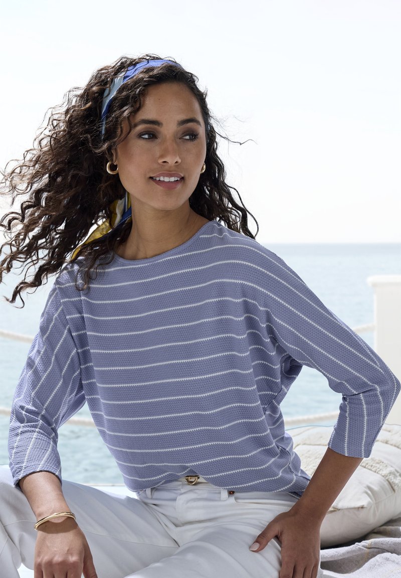 Frau mit lockigem Haar, die ein blau-weiß gestreiftes Oberteil, weiße Hose und ein blaues Kopftuch trägt und an einem sonnigen Tag am Meer sitzt.