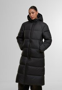Svart puffjacka med lång design, med huva, dragkedja fram och quiltat mönster. Materialet verkar slätt och isolerat.